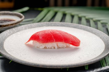 Minimalist bir tabakta Lean Tuna Maguro Nigiri. Parlak kırmızı bir maguro ton balığı parçası pirinçli beyaz dokuma bir tabakta duruyor. Etrafı çubuklarla çevrili ve koyu renkte..