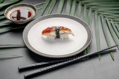Unagi Yılanbalığı Nigiri ve Susam Tohumu ve Karanlık Arkaplan. Susamlı ızgara unagi yılanbalığı nigiri, yanında koyu soya sosuyla minimalist bir seramik tabakta sunuluyor..