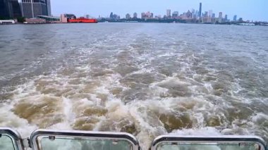 Uzak Şehir Skyline 'lı bir teknenin uyanışı. New York şehrinin uzak ufuk çizgisiyle çalkalanan köpüklü suyu gösteren bir teknenin arkasından manzara.