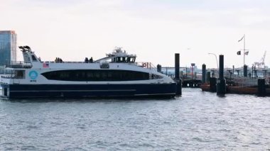 New York, ABD, 5 Aralık 2025: NYC Feribot Doğu Nehri rıhtımına yaklaşıyor. Mavi ve beyaz bir New York feribotu nehir kıyısındaki bir rıhtıma doğru ilerliyor. Güvertede yolcular ve soluk bir sanayi silueti var..