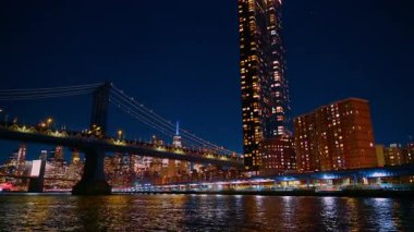 New York şehri geceleri mimarisini sergiliyor. Su, binaların ve köprünün ışıklarını yansıtıyor. Ufuk çizgisi arka planda görünür..