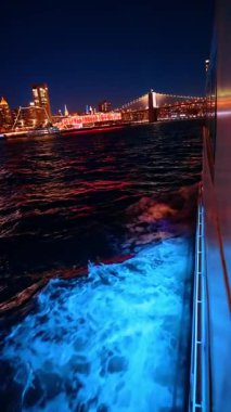 Şehir ışıkları yüzeyden yansıtırken bir tekne karanlık suda hareket ediyor. Manzara, New York 'ta geceleri yanan binalar ve bir köprüyü içeriyor..