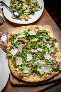 Tahta tahtada servis edilen taze malzemeli lezzetli pizza. Pizza yeşillik ve peynirle kaplıdır. Yanında da yemek masasında taze bir salata..