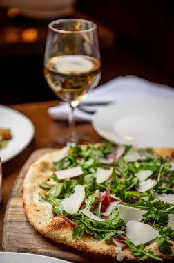 Restoran masasında beyaz şarabın yanında yeşil sebzeli taze pizza. Yeşillik ve peynirle kaplı bir pizza bir masada bir bardak beyaz şarabın yanında tahtanın üzerinde oturur..