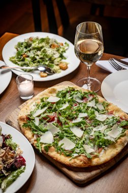 Samimi bir restoran ortamında salata ve şarapla servis edilen lezzetli pizza. Yeşilliklerle kaplı taze pizzayla dolu bir masa. Yanında salata ve bir bardak şarapla birlikte..