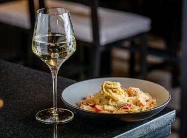 Akşam yemeği tabağında makarna ve siyah bir masada bir bardak beyaz şarap. Bir tabak makarna, akşam yemeği sırasında bir restoranda bir masada beyaz şarabın yanında oturur..