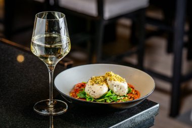 Restoran ortamında peynir, sebze ve bir bardak şarapla dolu bir tabak. Tabakta peynir, sebze ve fındık var. Restoranda beyaz şarap bardağının yanında..