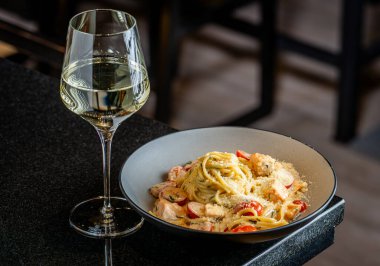 Yemek masasında bir bardak beyaz şarapla servis edilen lezzetli makarna tabağı. Domates ve peynirli bir tabak makarna bir restorandaki karanlık bir masada bir bardak beyaz şarabın yanında oturur..
