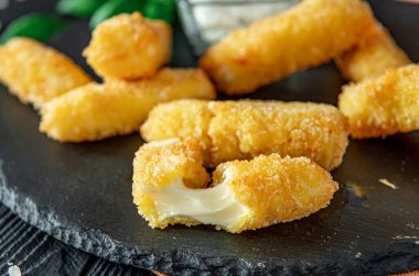 Koyu bir tabakta yeşil garnitürlü ve soslu peynir çubukları. Tabakta soslu ve yeşil garnitürlü kızarmış peynir çubukları var. Bazı peynir çubukları kırılmış..