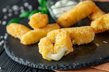 Mutfak masasında sosla servis edilen kızarmış peynir çubukları. Kızarmış peynir çubukları yanında küçük bir kase sosla birlikte siyah bir tabağın üzerinde. Altın ve çıtır çıtır..