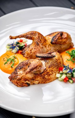 İki kızarmış bıldırcın ve beyaz tabakta renkli sebzeler. İki kavrulmuş bıldırcın, etraflarını saran bir sos ve sebze garnitürleriyle beyaz bir tabakta dinleniyor..