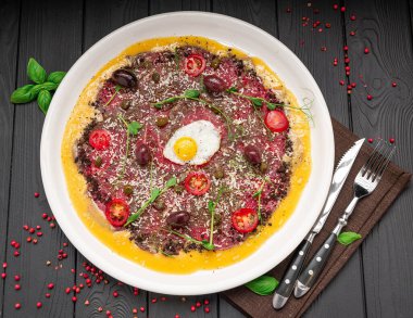 Restoranda beyaz bir tabakta yumurta ve sebzeli biftek carpaccio. Biftek carpaccio, basit dekorlu bir restoran masasında yumurta ve sebzelerle servis edilir..