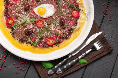 Koyu ahşap bir masada çiğ et, yumurta ve sebzeli lezzetli bir yemek. Koyu ahşap bir masa üzerinde yumurta, kiraz domatesleri ve otlarla kaplı çiğ et içeren bir yemek..