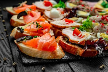 Koyu ahşap bir masada sergilenen lezzetli, açık yüzlü sandviçler. Farklı malzemelerle birçok açık yüzlü sandviç yemek sırasında siyah bir taş tepside servis edilir..