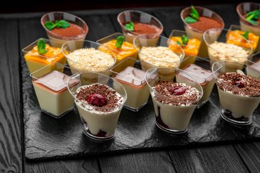 Koyu yüzeyde tatlı bardaklarında krema, meyve ve çikolata katmanları. Krema, meyve ve çikolatayla dolu çoklu tatlı fincanları koyu bir taş yüzeyde oturur..