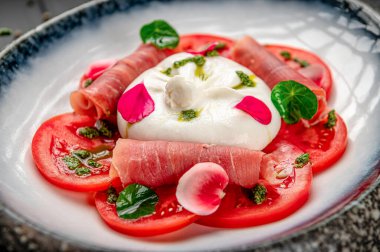 Taze domates ve jambonlu burrata peyniri ve tabakta otlar. Taze domatesli bir tabak, burrata peyniri, prosciutto ve en üste konmuş otlar..