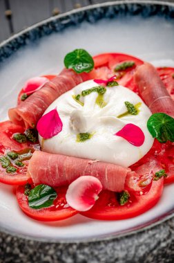 Peynirli taze salata, prosciutto ve bir tabağa konmuş şifalı bitkiler. Bir kasede dilimlenmiş domates, krem peynir, prosciutto ve çiçek yapraklı otlar bulunur..