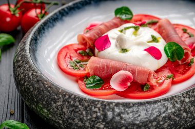Koyu ahşap bir masada burrata peynirli ve jambonlu taze domates salatası. Bir salata dilimlenmiş domates, burrata peyniri ve yeşil bir tabakta hazırlanmış jambon içerir..