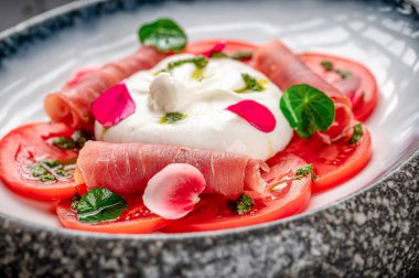 Bir kasede servis edilen domatesli, jambonlu ve peynirli taze salata. Jambon, peynir ve otlarla kaplı taze domates dilimleri bir kasede duruyor..