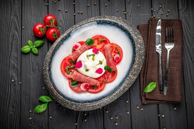 Taze domates ve mozzarella peyniri. Koyu ahşap bir masada, çatal bıçaklarla. Dilimlenmiş domates ve mozzarella peyniri bir tabakta otlarla ve et dilimleriyle sıralanmış..