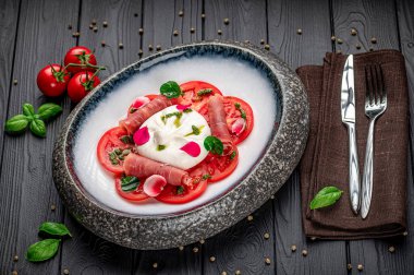 Koyu ahşap bir masada çatal bıçakla servis edilen taze domates ve peynir tabağı. Dilimlenmiş domates ve peynirli bir tabak karanlık bir masaya konur..