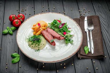 Chef prepares a meal with meat, vegetables, and salad on a table. A plate holds cooked meat, mashed potatoes, salad, and fresh tomatoes beside cutlery on a wooden table.