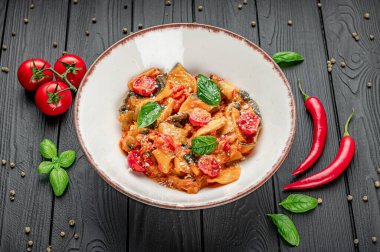 Tahta masada domatesli ve fesleğenli taze makarna tabağı. Bir kase domatesli makarna, fesleğen ve kırmızı biber taze malzemelerle çevrili ahşap bir masaya konur..