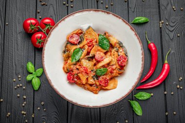 Domatesli makarna, biber ve ahşap bir masada fesleğen. Etrafına domates, acı biber ve fesleğen konmuş koyu ahşap bir masaya bir kase makarna konur..