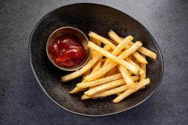 Kızartmalar bir kasede, koyu bir yüzeyde ketçap ile servis edilir. Bir kase, üzerinde kırmızı ketçap bulunan küçük bir fincanın yanında altın patates kızartması içerir..