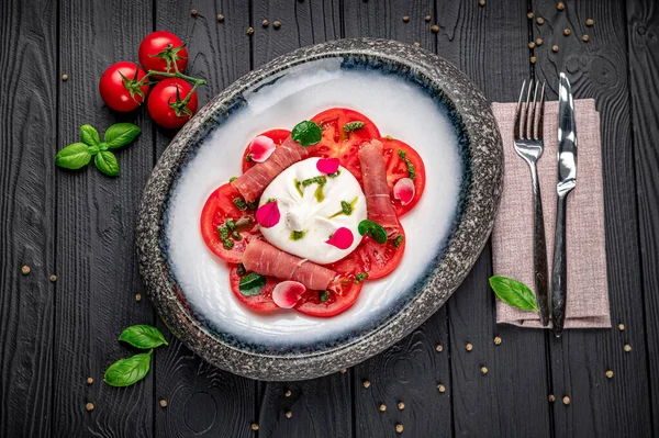 Taze domates ve peynir bir tabakta otlar ve sebzelerle servis edilir. Bir tabak taze domates, peynir, otlar ve ahşap bir masada kiraz domatesleri yer alır..