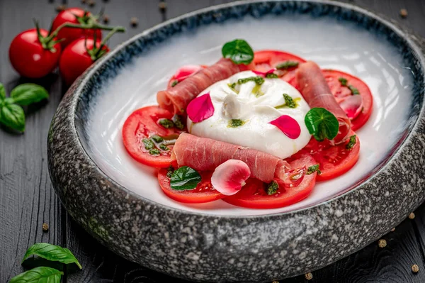 Domatesli salata, mozzarella ve siyah ahşap bir masada jambon. Bir tabak içinde domates, mozzarella topu ve jambon dilimleri var..