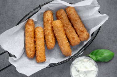 Gri bir yüzeyde soslu bir sepette servis edilen çıtır çerezler. Bir sepete koyulmuş altın kahverengi atıştırmalıklar, yanında sos ve yanında bitki yaprağı..