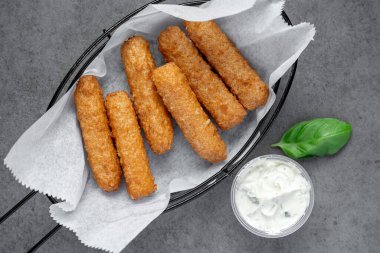 Gri bir masada sosla servis edilen çıtır çubuklar. Çıtır çubuklar, gri bir yüzeyde soslu siyah bir sepette düzenlenir..