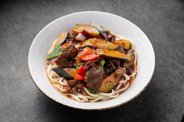 Basit bir tabakta servis edilen sığır eti ve sebzeli sıcak erişte kasesi. Biftek ve sebzeyle kaplanmış erişteler karanlık bir yüzey üzerinde bir kasede servis edilir..