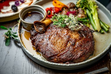 Steak with vegetables and sauce served in a restaurant setting. A large piece of steak is served with grilled vegetables and sauce on a plate at a dining table.