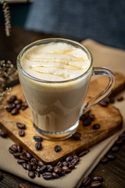 Kahve içeceği, kahve çekirdekleri ve bezlerle çevrili bir tahtanın üzerinde duruyor. Köpüklü sütlü bir fincan kahve ahşap bir tahtanın üzerinde dağınık kahve çekirdekleri ile oturur..