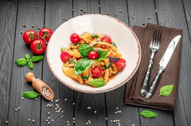 Kiraz domatesli taze makarna tabağı ve ahşap bir masada fesleğen. Bir kase taze makarna, yakınlardaki kiraz domatesli, fesleğenli ve gümüş çatal bıçaklı ahşap bir masada duruyor..