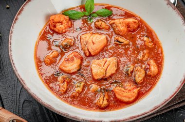 Tahta bir masada taze otlarla servis edilen deniz ürünleri güveci. Bir kase, karanlık bir masada karides, balık ve kabuklu deniz ürünleri güveci içerir..