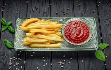 Ketçapla servis edilen patates kızartması ve karanlık bir masada taze otlar. Patates kızartması bir tabak ketçap ve karanlık bir masanın üzerine serpilmiş otların yanına konur..