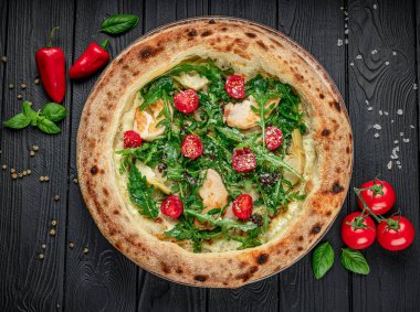 Tahta masadaki taze malzemelerle nefis bir pizza. Yuvarlak bir pizza ahşap bir masada oturur. Yanında tavuk, domates ve biberli roka gibi malzemeler var..