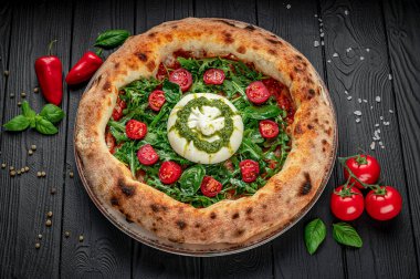 Domatesli taze pizza ve ahşap bir masada fesleğen. Taze domates, fesleğen ve kremalı peynirle kaplı yuvarlak bir pizza koyu ahşap bir masada oturur..