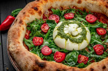 Fesleğenli taze pizza, domates ve siyah ahşap bir masada kremalı peynir. Lezzetli bir pizza, bitkilerle, domateslerle ve krem peynirle kaplı bir masada oturur..