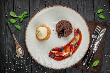 Beyaz bir tabakta taze meyve ve dondurmayla servis edilen lezzetli bir tatlı. Bir tatlı tabağında yeşil yapraklı bir masa üzerinde çikolatalı pasta, dondurma, meyve ve sos yer alır..