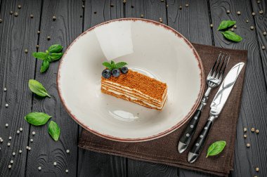 Sofrada çatal bıçak takımı ve fesleğen yapraklarıyla servis edilen tatlı. Bir tatlı parçası beyaz bir tabağa, çatal bıçak takımının yanına ve ahşap bir masada taze fesleğen yapraklarının yanına konur..