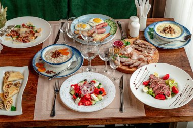 Çeşitli yemekler ve ahşap bir yemek masasında içeceklerle nefis bir yemek. Tahta bir masaya farklı tabaklar koyulmuş. Yanında da bardaklar ve çatal bıçak takımı var..