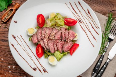 Dilimlenmiş sığır eti, salata ve ahşap bir masada sosla servis edilir. Dilimlenmiş et, domatesli ve yumurtalı bir salata yatağına konur.