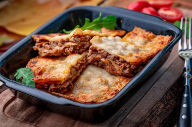 Tahta bir masaya konmuş siyah bir konteynırda lazanya ve yanında domatesler. Siyah bir kap, yanında taze domateslerle ahşap bir masada etli ve peynirli lazanya katmanları barındırır..