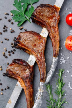 Gümüş bir rafta ızgara kuzu pirzolası ve baharat. Üç ızgara kuzu pirzolası gümüş bir rafa konur ve etraflarında gri bir yüzey üzerinde otlar, biberler ve domatesler bulunur..