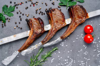 Gri bir masada baharatlı ve otlu ızgara kuzu pirzolası. Izgara kuzu pirzolası metal bir rafta duruyor. Gri bir yüzeye yakın otlar, baharatlar ve taze domateslerle birlikte..