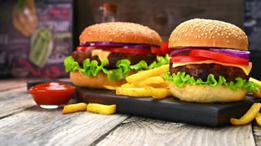 Tahta bir masada patates kızartması ve ketçaplı iki hamburger. İki hamburger, marul, domates ve soğanla ahşap bir tahtanın üzerinde patates kızartması ve küçük bir kase ketçapın yanında oturur..